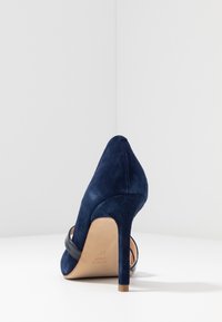 Scarpa con tacco alto in suede blu navy, con punta affusolata e tacco sottile, fotografata da dietro su una superficie bianca.