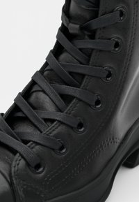 Zwart lederen enkellaars met zes paar zwarte oogjes, platte zwarte veters en een gladde textuur. Stikseldetail zichtbaar langs de zijkant.