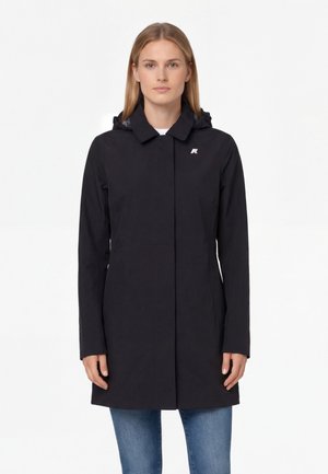 Femme portant un imperméable noir à capuche avec un petit logo sur la poitrine, debout droite devant un fond blanc uni.