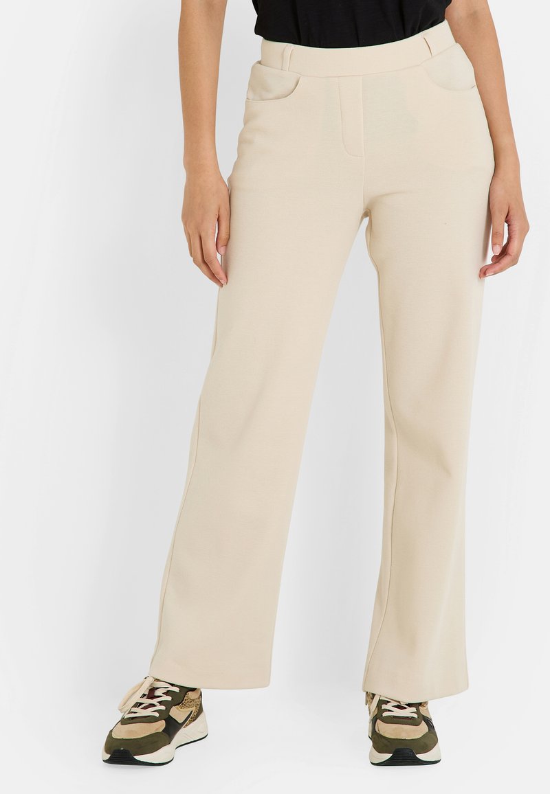 Pantalones de pierna ancha color beige, hechos de un tejido suave, con dos bolsillos frontales y una cinturilla lisa, combinados con zapatillas negras y verdes.