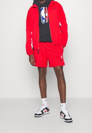 Osoba w czerwonej kurtce i szortach Nike, czarnej bluzie NBA, białych skarpetkach Nike oraz czarno-biało-czerwonych butach sportowych Nike stoi na tle białej ściany.