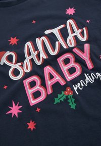 Temno modra majica s sijočim in barvitim napisom "SANTA BABY", okrašena s trtnimi listi in roza zvezdicami kot dekorativnimi elementi.