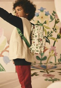 Bílý látkový batoh s barevným květinovým a karakterovým potiskem, dvěma zipovými kapsami, zelenými popruhy, kombinovaný s krémovou vestou a červenými kalhotami.