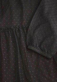 Czarna siatkowa tkanina z małymi, rozproszonymi kropkami. Posiada marszczone detale w talii oraz elastyczne mankiety przy balonowych rękawach.