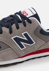 Grå mockaskor med marinblå nätakcenter, med en vit och marinblå "N"-logotyp, röd detalj på mittsålen och mörk gummisul.