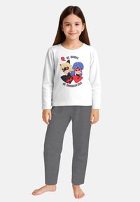 Young girl with long brown hair wearing a white long-sleeve shirt featuring two animated superheroes and the text "Be Brave Be Miraculous."