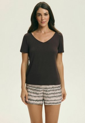 Kobieta z długimi ciemnymi włosami, ubrana w czarną koszulkę z krótkim rękawem o dekolcie w serek oraz czarno-białe szorty w wzory, stojąca przodem.