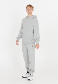 Ung man som står och ler, klädd i ljusgrå hoodie och matchande sweatpants med vita sneakers, på en enfärgad vit bakgrund.