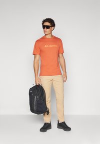 Oransje kortermet t-skjorte med Columbia-logo, beige bukser, svart ryggsekk med glidelåser, og svarte sko. Glatt stoff og moderne design.
