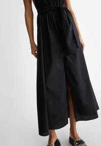 Schwarzes Maxi-Kleid mit elastischem Bund und Gürtel, ausgestattet mit einem seitlichen Schlitz und glatter Stofftextur, kombiniert mit Schuhen im Tiermuster.