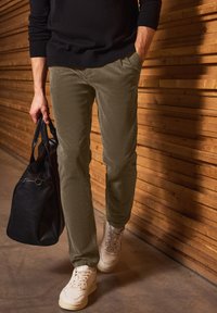 Jasnooliwkowe spodnie wykonane z miękkiego materiału, połączone z białymi sneakersami. Czarna torba trzymana jest w jednej ręce na tle drewnianym.