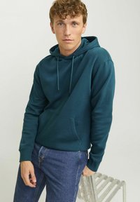 Tealfarbene Kapuzenjacke aus weichem Stoff mit einer Fronttasche, verstellbaren Kordeln und gerippten Bündchen. Kombiniert mit blauen Jeans.