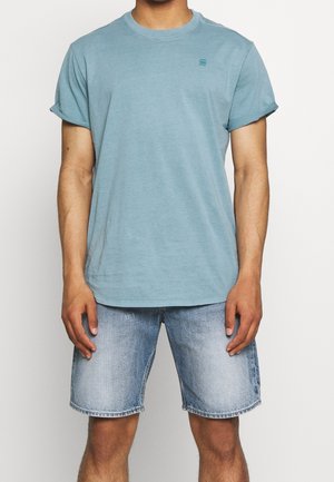 Mężczyzna w jasnoniebieskim t-shircie z podwiniętymi rękawami oraz wyblakłych niebieskich szortach jeansowych stoi na białym tle.