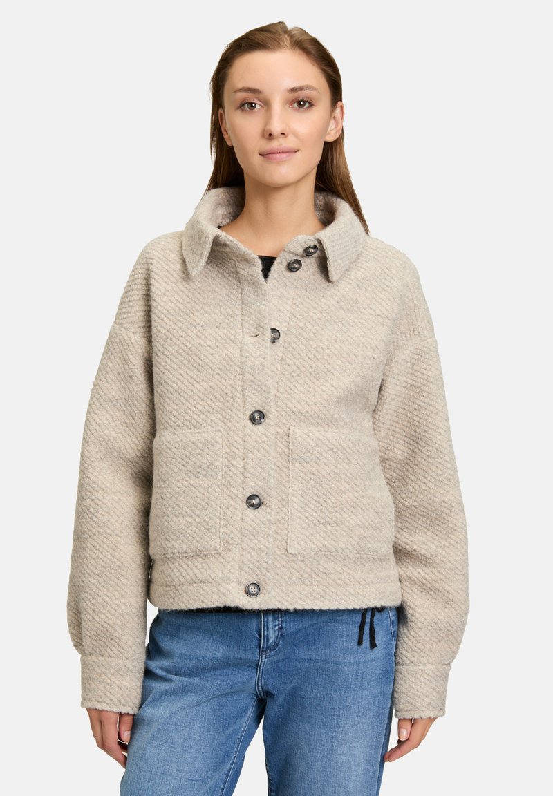 Veste cropped beige en tissu texturé, avec un grand col, quatre boutons à l'avant et deux poches sur le devant, associée à un jean bleu.