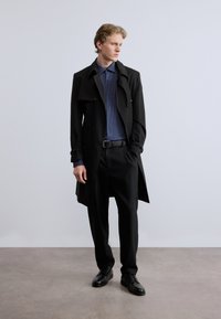 Μαύρο παλτό τρανς πάνω από ένα σκούρο μπλε πουκάμισο με πουά, μαύρο παντελόνι και μαύρα παπούτσια· κομψό σχέδιο με κούμπωμα κουμπιών και γυρισμένα μανίκια.