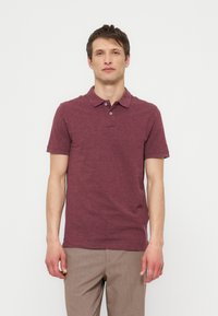 Bărbat tânăr în picioare cu fața spre înainte, purtând un tricou polo burgund cu mâneci scurte și pantaloni maro deschis pe un fundal alb simplu.