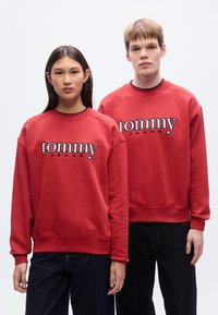 Κόκκινα φούτερ με το λογότυπο «tommy», κατασκευασμένα από βαμβακερό μείγμα, με ριμπ μανσέτες και σχέδιο με στρογγυλή λαιμόκοψη, σε συνδυασμό με σκούρο τζιν παντελόνι.