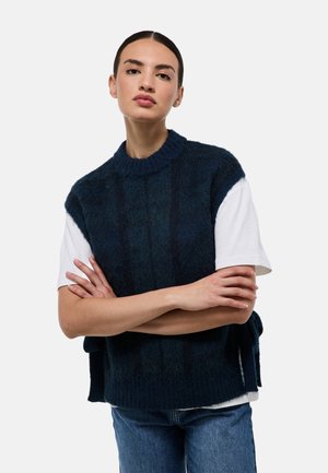 Haut sans manches en tricot bleu marine avec un motif texturé, un col rond et une coupe ample, superposé à une chemise blanche à manches courtes.