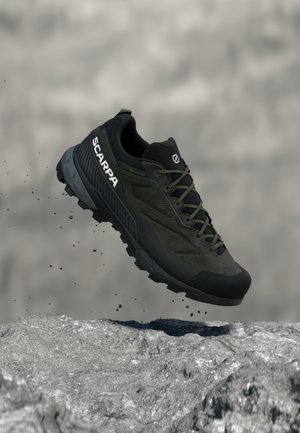 Zapato de senderismo Scarpa negro y verde con suela resistente suspendido sobre una superficie rocosa contra un fondo desenfocado.