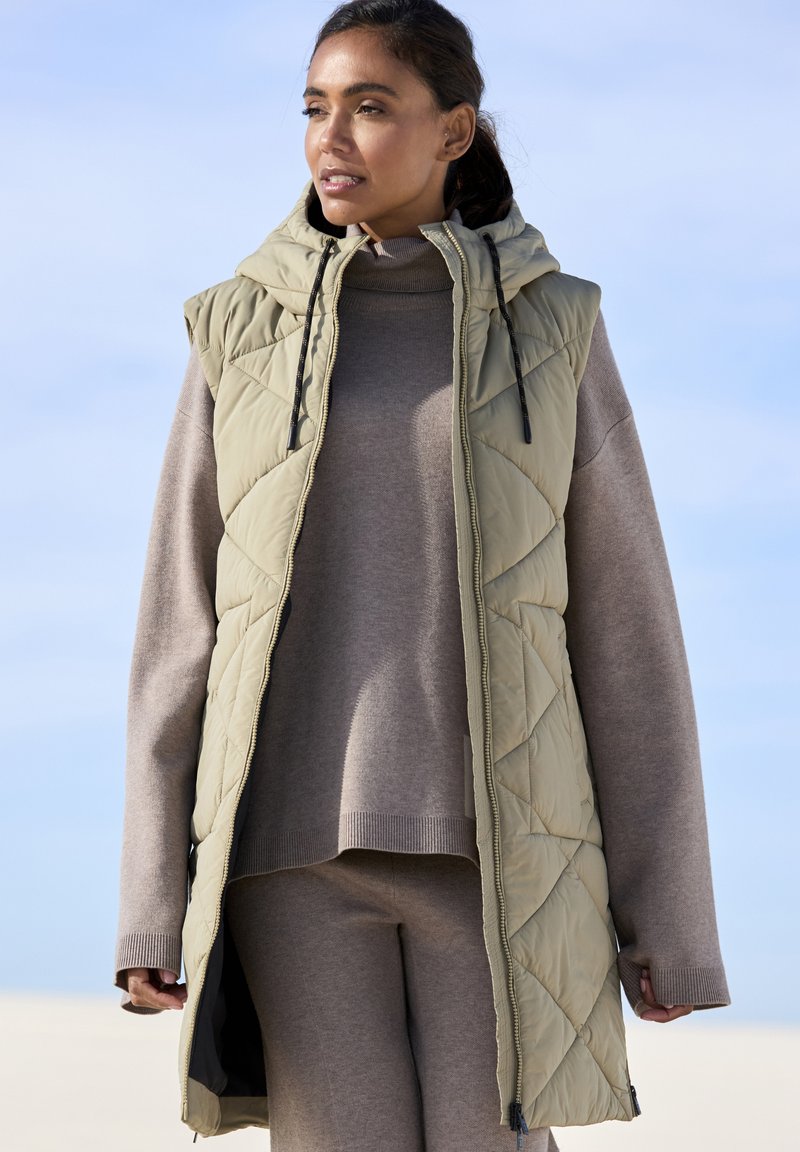 Khaki gesteppte Weste mit Kapuze, durchgehendem Reißverschluss und Seitentaschen, über einem beigen Pullover und passenden Hosen getragen. Glatte Textur.