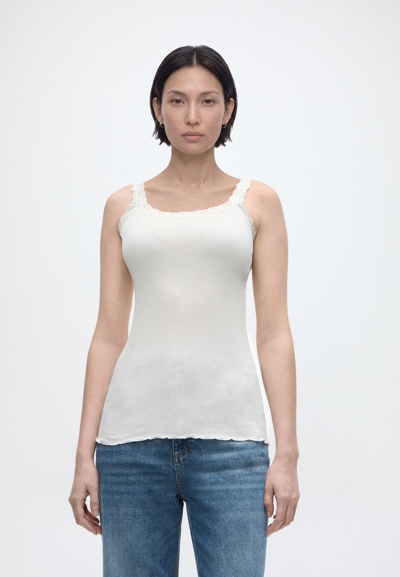 Γυναίκα με ίσια σκούρα μαλλιά φοράει λευκό ριμπ αμάνικο τοπ με δαντέλα και μπλε τζιν, στέκεται μπροστά σε απλό ανοιχτό φόντο.