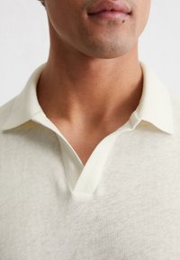 Light cream polo shirt with a V-neck collar. Made of textured fabric, featuring short sleeves and a subtle, soft finish.