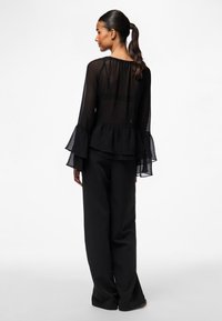 Black, sheer blouse with ruffle details on the sleeves and a peplum hem, paired with black wide-leg trousers. Smooth fabric texture.