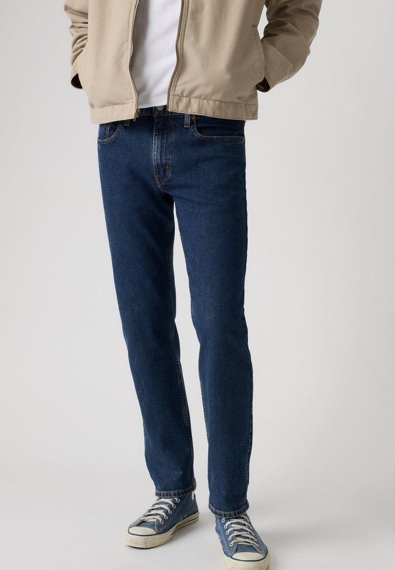 Homme portant un jean bleu, des baskets en toile bleues, un t-shirt blanc et une veste beige zippée, debout les mains dans les poches.