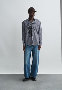 Svītrains garām piedurknēm krekls ar melnu grafiku, brīvi pieguļoši zili denima džinsi un melnas kurpes, papildināti ar brūnu cepuri.