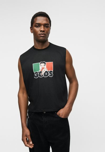 Άνδρας που φοράει μαύρο αμάνικο μπλουζάκι με καρτούν χαρακτήρα και το κείμενο "GCDS", στέκεται με το ένα χέρι στην τσέπη μπροστά σε απλό φόντο.