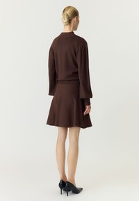 Kvinde med blond hår i en knold iført en brun langærmet sweater og matchende nederdel, stående med ryggen mod fronten, iført sorte høje hæle.