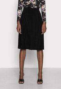 Jupe plissée noire avec une taille haute, texture douce, longueur genou, associée à des chaussures noires à bout ouvert. Le haut présente un motif floral.