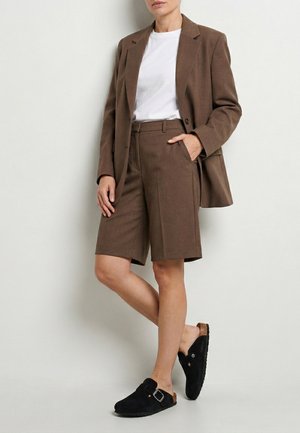 Kvinde iført brun blazer og matchende shorts til knælængde, hvid t-shirt og sorte slip-on sko med spænde, der står op ad en hvid væg.