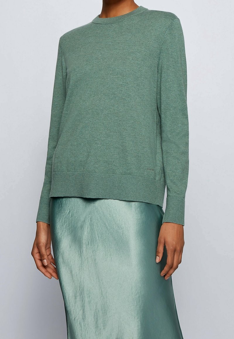 Personne portant un pull en tricot vert sauge à manches longues et une jupe midi en satin brillant assortie, debout devant un fond gris clair.