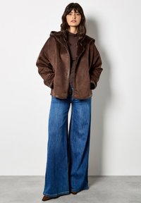 Giacca in finta pelliccia shearling marrone con cappuccio, caratterizzata da una chiusura a pulsanti, vestibilità oversize e tasche profonde, abbinata a jeans in denim blu a gamba larga.