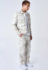 Chaqueta de mezclilla de color claro con múltiples bolsillos, textura con patrones, combinada con pantalones cargo a juego. El modelo se encuentra de pie frente a un fondo liso.