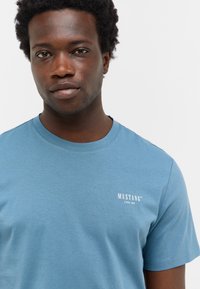 Niebieska koszulka z krótkim rękawem, wykonana z miękkiego materiału, z okrągłym dekoltem i białym nadrukiem logo "MUSTANG" na lewej piersi.