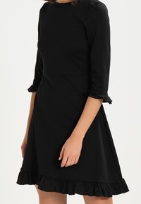 Robe noire avec manches trois-quarts, poignets à volants et ourlet évasé. Fabriquée en tissu lisse, avec un corsage ajusté.