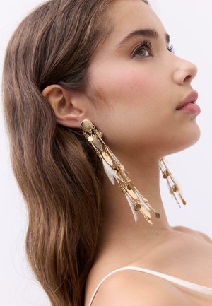 Giovane donna con capelli castani lunghi, che indossa orecchini a catena lunga in oro con piccoli dischi e dettagli in piuma bianca, che guarda verso l'alto.