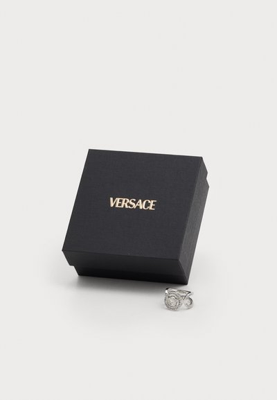 Versace ring in silver with circular stone and accent stones, placed beside a black textured box with gold lettering.