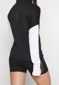 Musta urheilu paita valkoisilla hihoilla, jossa on korkea kaulus, tyylikäs muotoilu ja takana pieniä valkoisia pisteitä. Yhdistetty mustiin shortseihin.