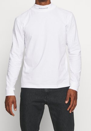 Mężczyzna noszący gładką białą koszulę z długim rękawem z napisem "Calvin Klein Jeans" na kołnierzu oraz ciemnoszare dżinsy.