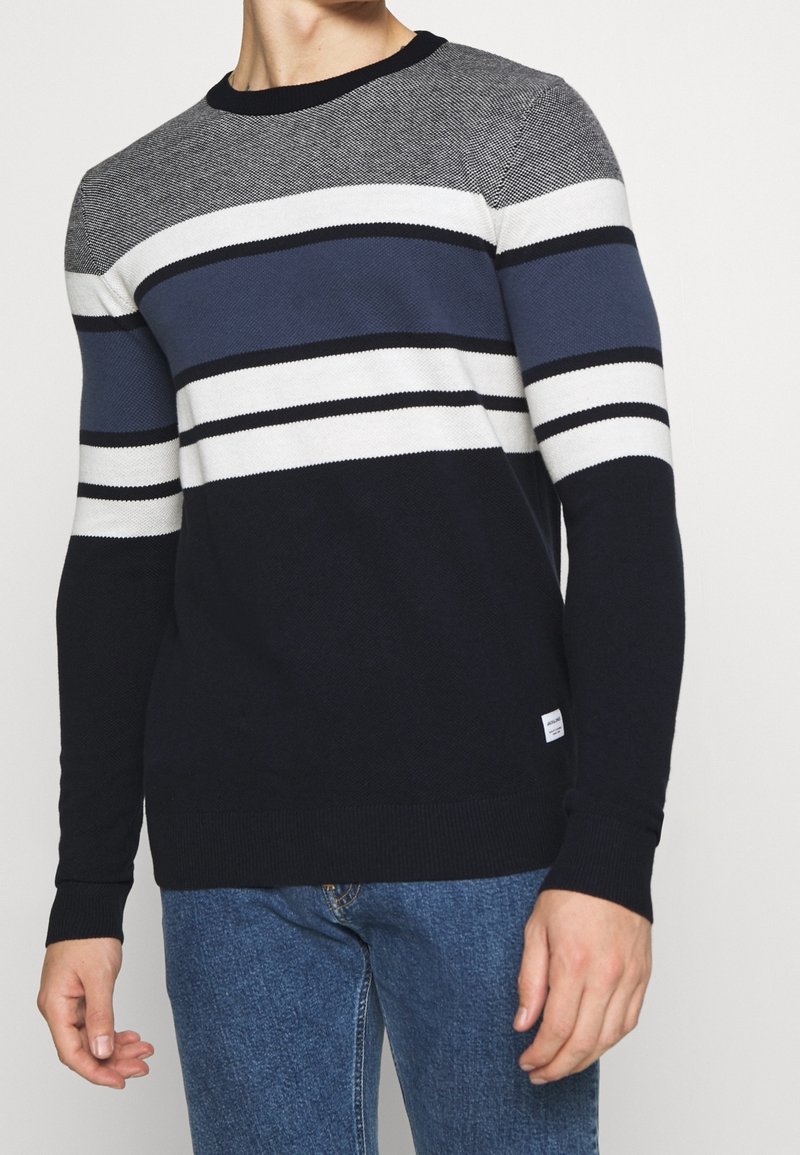 Homme portant un pull en maille à manches longues avec des rayures horizontales noires, blanches, grises et bleues, assorti à un jean bleu.