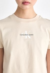Bežinė medvilninė marškinė su apvalia iškirpte. Ant jų yra pilkšvai baltas užrašas "Calvin Klein Jeans". Trumpomis rankovėmis, lygaus tekstūros.