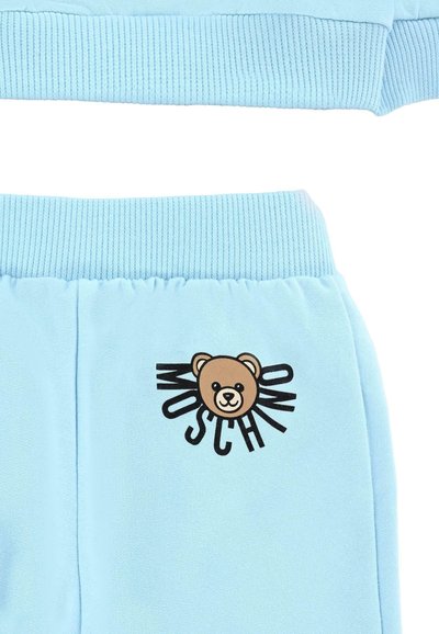 Pantaloni della tuta azzurro chiaro con vita a costine, caratterizzati da una grafica di un orso bruno e il testo “MOSCHINO” in nero. Tessuto morbido con una texture liscia.