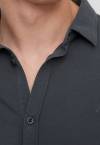 Dunkelgraues Polohemd mit strukturiertem Stoff, kurzem Kragen und Knopfverschluss. Verfügt über ein kleines Logo auf der linken Seite oberhalb des Knopfes.