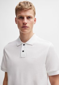 Bílá polo košile vyrobená z bavlny, s límečkem, tříknoflíkovou légu s černými knoflíky a decentním vyšitým logem BOSS na hrudi.