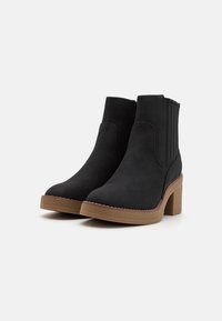 Bottes chelsea en daim noir avec un talon épais et texturé, des panneaux latéraux nervurés et une semelle contrastante en marron. Design classique avec des lignes épurées.