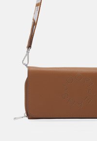 Brązowa skórzana saszetka na nadgarstek z perforowanym wzorem logo, zamknięciem na suwak oraz kontrastowym paskiem z wzorem przymocowanym metalowym zaczepem.