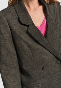 Blazer de doble botonadura en gris oscuro, llevado sobre un top de cuello en V rosa brillante, que muestra la parte superior del torso y los detalles del collar.